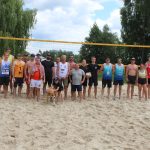 Na zdjęciu widzimy wszystkich uczestników turnieju piłki siatkowej plażowej nad Jeziorem Białym stojących na boisku do siatkówki plażowej.