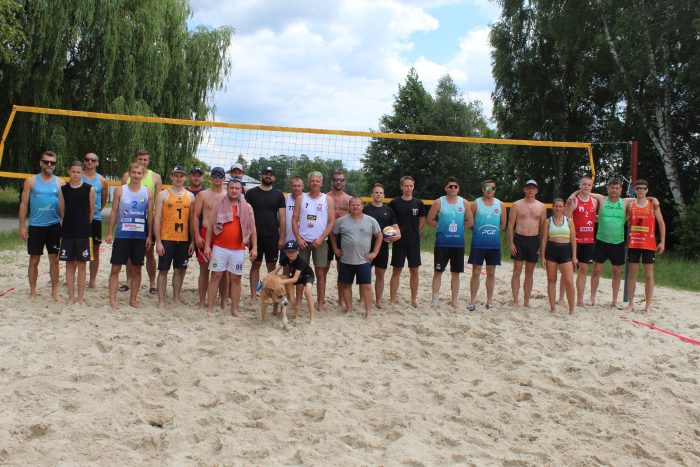 Na zdjęciu widzimy wszystkich uczestników turnieju piłki siatkowej plażowej nad Jeziorem Białym stojących na boisku do siatkówki plażowej.