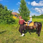 Na zdjęciu widzimy dziecko na koniu oraz pracownika obsługi na błoniach przy Kiszarni w Orchówku.