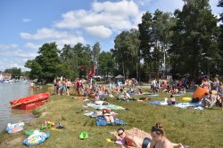 Na brzegu jeziora leży mnóstwo ludzi w strojach kąpielowych. W oddali widać wieżyczkę z ratownikiem, a w tle zielone drzewa.