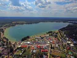 Jezioro Białe z lotu ptaka – Widok z lotu ptaka na niebieskie jezioro z mnóstwem kąpiących się ludzi, otoczone z jednej strony gęstą zabudową, z drugiej lasem.