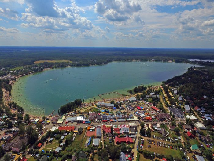 Jezioro Białe z lotu ptaka – Widok z lotu ptaka na niebieskie jezioro z mnóstwem kąpiących się ludzi, otoczone z jednej strony gęstą zabudową, z drugiej lasem.