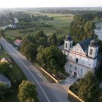 Kościół pw. św. Jana Jałmużnika w Orchówku – Widok z lotu ptaka na biały kościół z dwiema wieżami oraz na szosę i zabudowania przy niej. W tle widać drzewa oraz porośnięty rzęsą zbiornik wodny.