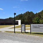 Muzeum i Miejsce Pamięci w Sobiborze – Widok na nowoczesny budynek o elewacji z paneli, do którego wiodą alejki wysypane jasnym żwirem. Z prawej strony stoi latarnia oraz tablica informacyjna, dalej widać las.