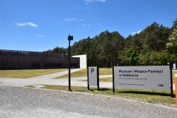 Muzeum i Miejsce Pamięci w Sobiborze – Widok na nowoczesny budynek o elewacji z paneli, do którego wiodą alejki wysypane jasnym żwirem. Z prawej strony stoi latarnia oraz tablica informacyjna, dalej widać las.