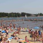Sezon wakacyjny w Okunince – Plaża z mnóstwem ludzi w strojach kąpielowych, na dalszym planie kąpielisko otoczone pomostem oraz jezioro z pływającą żaglówką.