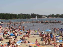 Sezon wakacyjny w Okunince – Plaża z mnóstwem ludzi w strojach kąpielowych, na dalszym planie kąpielisko otoczone pomostem oraz jezioro z pływającą żaglówką.