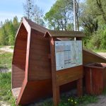 Tuba do słuchania odgłosów natury przy Trójstyku – Drewniana konstrukcja w kształcie tuby ustawiona przy gruntowej drodze. Obok widać drewniane tablicę informacyjną oraz kosz na śmieci.
