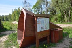 Tuba do słuchania odgłosów natury przy Trójstyku – Drewniana konstrukcja w kształcie tuby ustawiona przy gruntowej drodze. Obok widać drewniane tablicę informacyjną oraz kosz na śmieci.