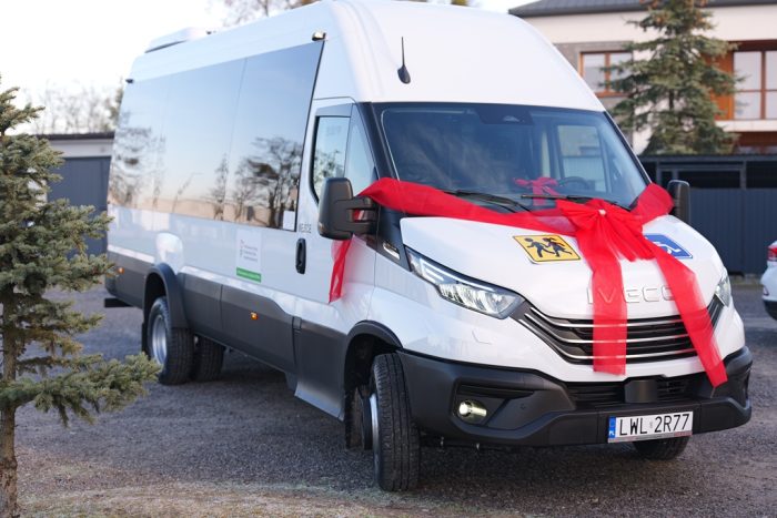 Biały bus marki Iveco stoi na parkingu. Na przedniej mace jest wielk czerwona kokarda