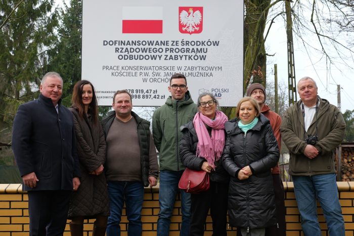 Na zdjęciu widać grupę osób pozujących na tle drzew i tablicy informacyjnej z nazwa projektu i kwotą jego dofinansowania